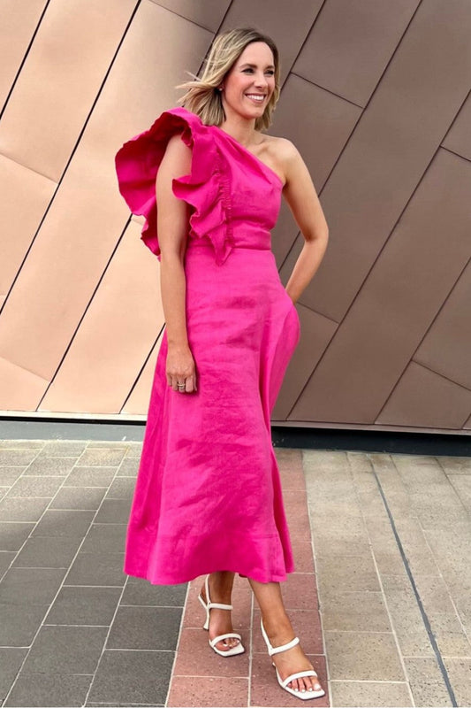 Woman in a bright pink one-shoulder Aje dress standing against a modern building.